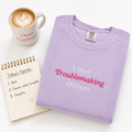 A folded lavender crewneck sweatshirt with 'Chief Troublemaking Officer' embroidered in white and pink lettering, styled on a white background with a coffee mug and a spiral agenda notebook.