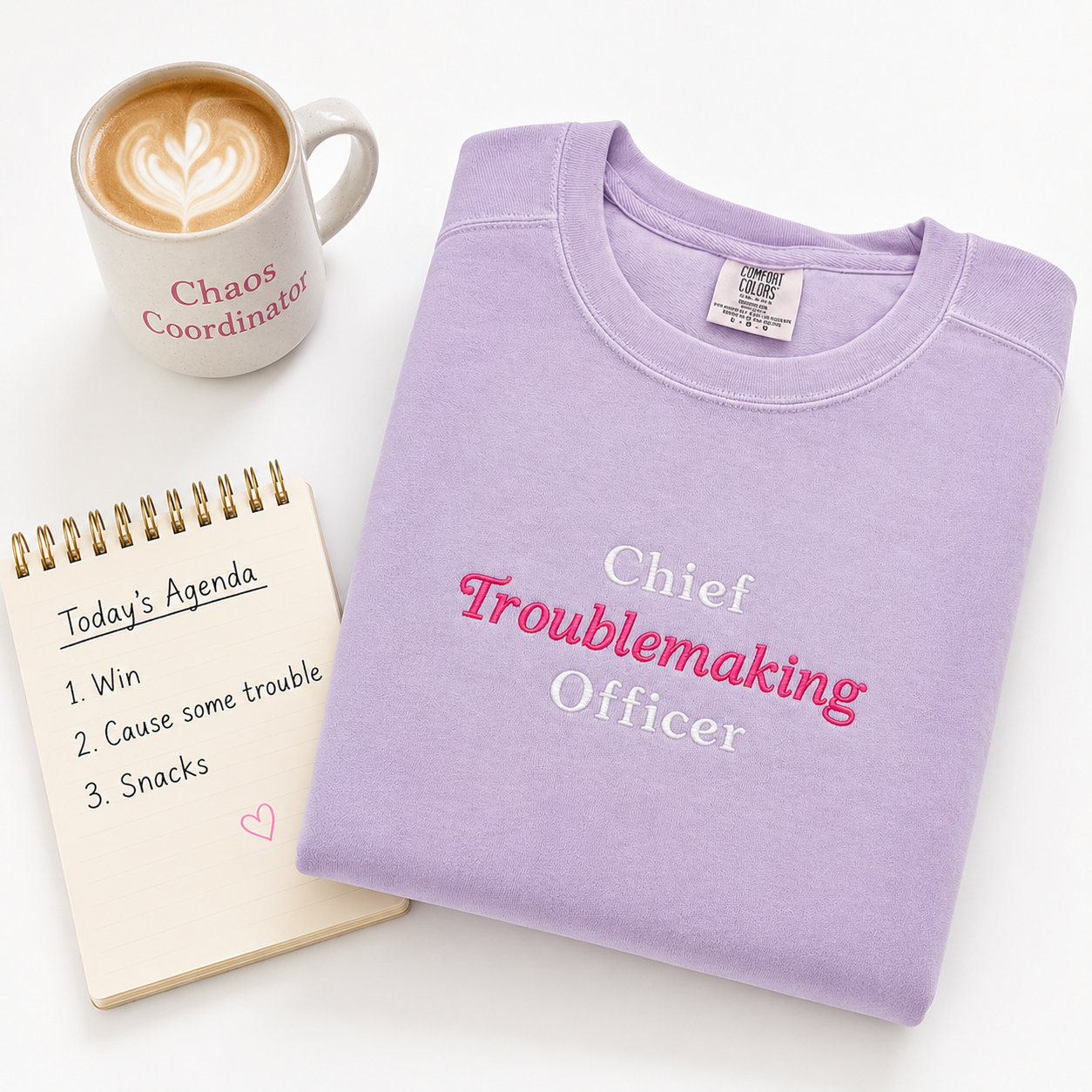 A folded lavender crewneck sweatshirt with 'Chief Troublemaking Officer' embroidered in white and pink lettering, styled on a white background with a coffee mug and a spiral agenda notebook.