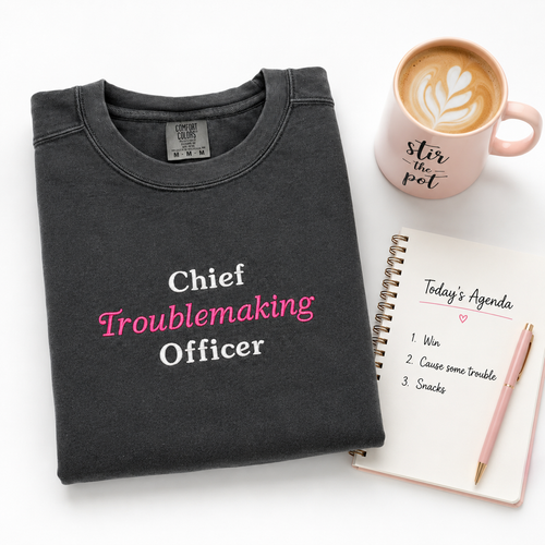 A folded dark charcoal gray t-shirt with the text 'Chief Troublemaking Officer' embroidered in white and pink lettering, styled in a flat-lay arrangement with a pink coffee mug, a spiral notebook, and a pen on a white background.