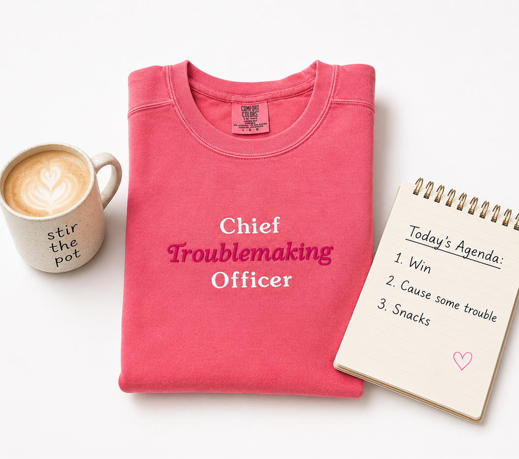 A folded pink crewneck sweatshirt with the text 'Chief Troublemaking Officer' embroidered in white and magenta, styled on a white background with a coffee mug and a notepad.