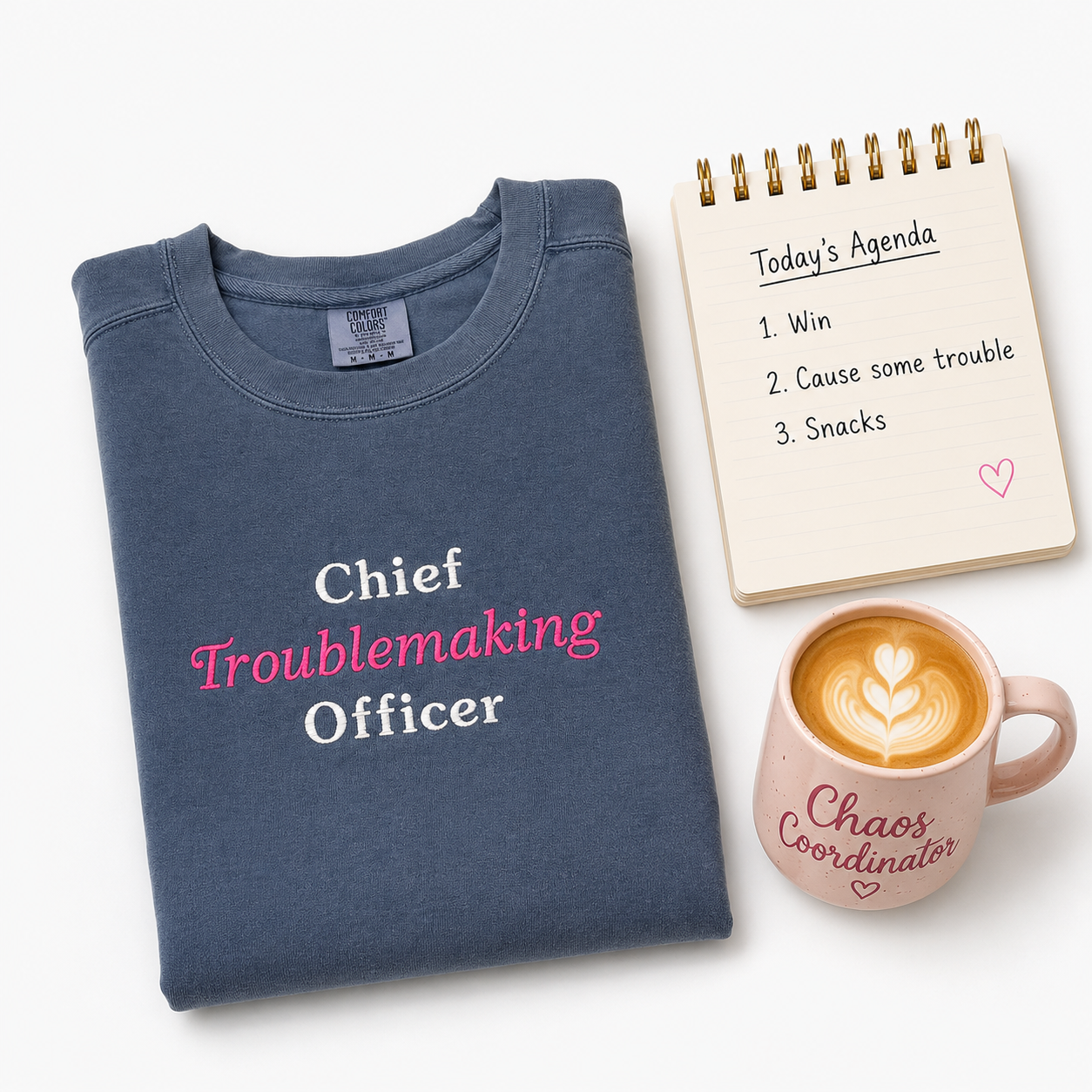 A folded navy crewneck sweatshirt with the text 'Chief Troublemaking Officer' embroidered in white and pink lettering, styled on a white background alongside a spiral agenda and a pink coffee mug.