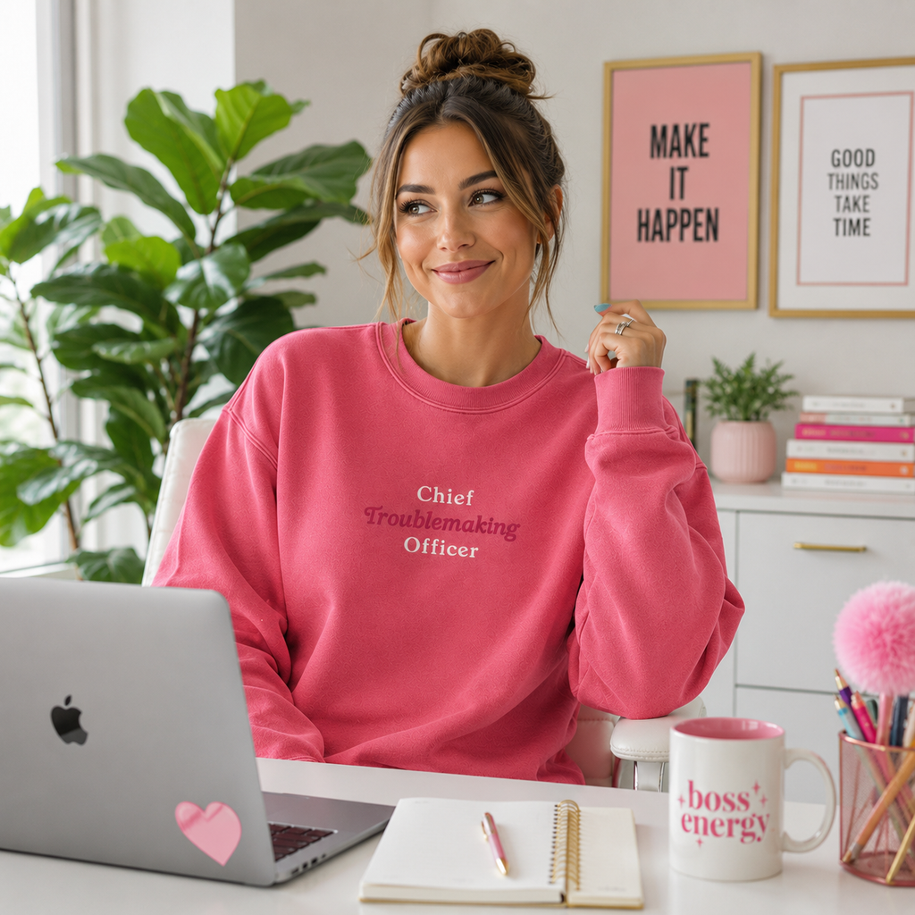 A woman sitting at a desk wearing a pink crewneck sweatshirt with the text 'Chief Troublemaking Officer' in white script. The scene is a bright, modern home office with a laptop, mug, plants, and framed wall art.