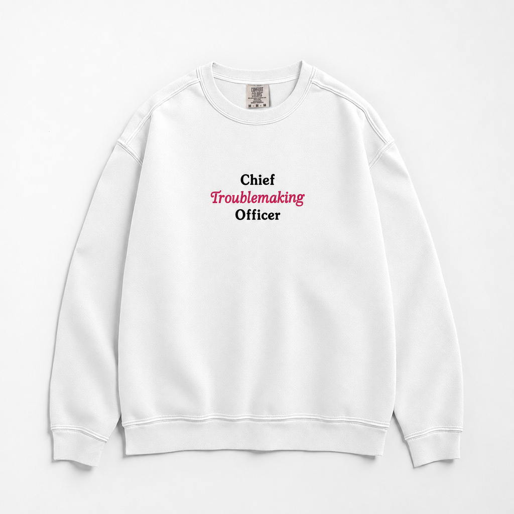 A white crewneck sweatshirt with the text 'Chief Troublemaking Officer' printed on the chest, featuring 'Chief' and 'Officer' in black and 'Troublemaking' in pink. The sweatshirt is laid flat against a clean white background.