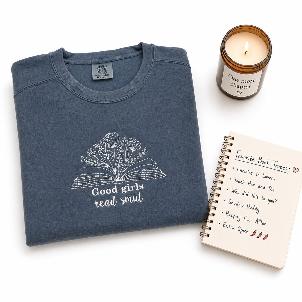 A folded slate blue crewneck sweatshirt with a white embroidered graphic of an open book and the text 'Good girls read smut' is displayed on a white background alongside a lit candle and a spiral notebook.