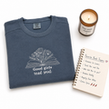 A folded slate blue crewneck sweatshirt with a white embroidered graphic of an open book and the text 'Good girls read smut' is displayed on a white background alongside a lit candle and a spiral notebook.
