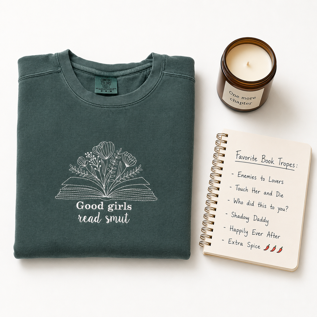 A folded dark green sweater with a white embroidered graphic of an open book made of flowers and the text 'Good girls read smut' is styled on a white background alongside a brown candle jar and a spiral notebook.