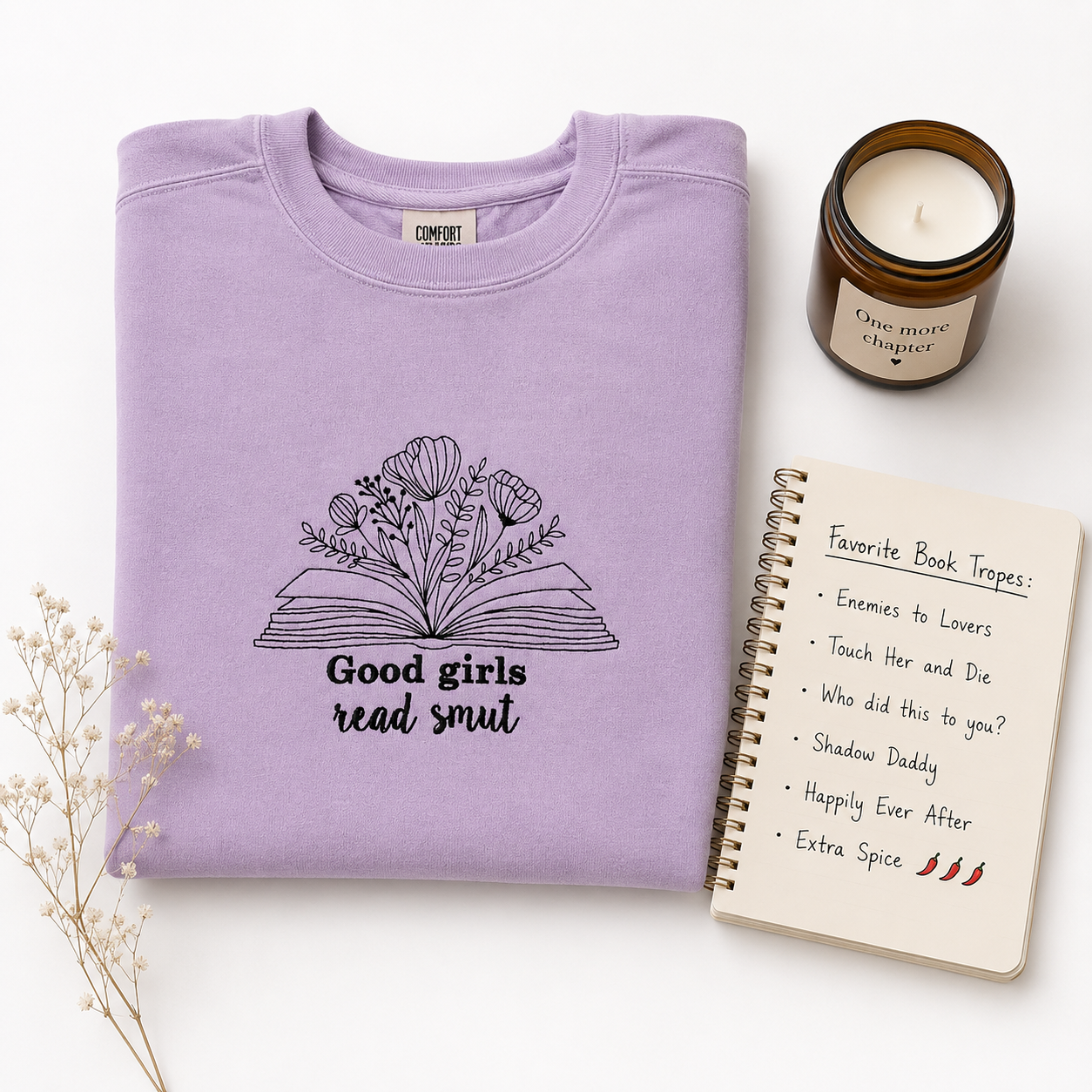 A folded lavender crewneck sweatshirt with a black embroidered graphic of an open book made of flowers and the text 'Good girls read smut'. The sweatshirt is styled in a flat-lay arrangement with a brown candle, a spiral notebook, and dried flowers on a white background.