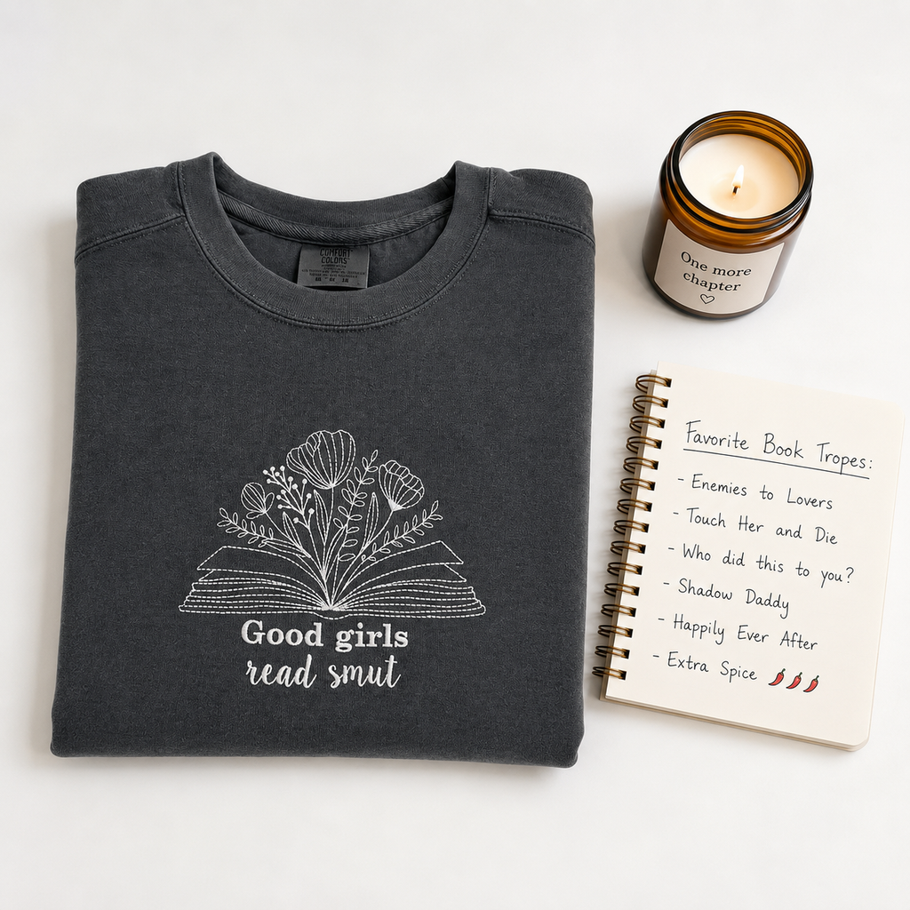 A folded dark charcoal gray t-shirt with a white embroidered graphic of an open book made of flowers and the text 'Good girls read smut' is styled on a white background next to a lit amber glass candle and a spiral notebook.