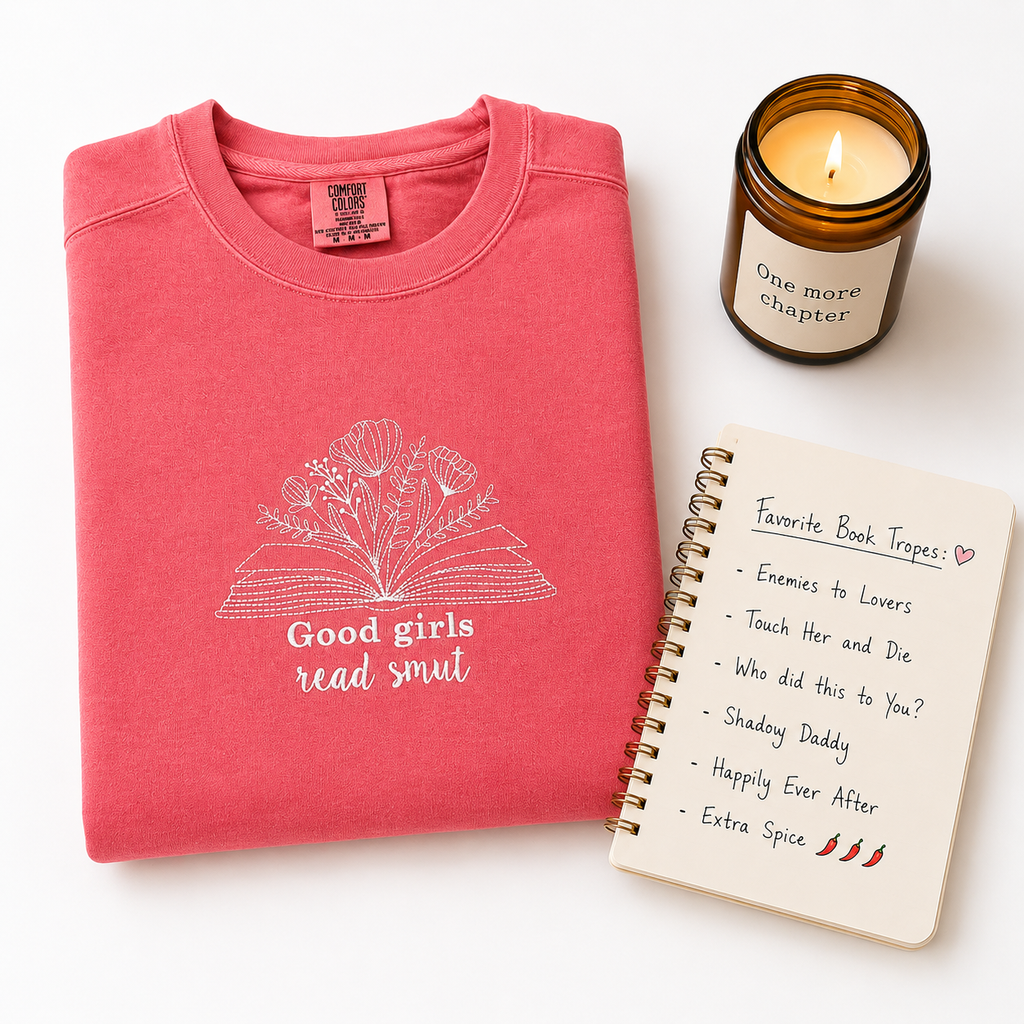 A folded coral pink crewneck sweatshirt with a white embroidered graphic of an open book and the text 'Good girls read smut' is displayed on a white background alongside a lit amber glass candle and a spiral notebook.