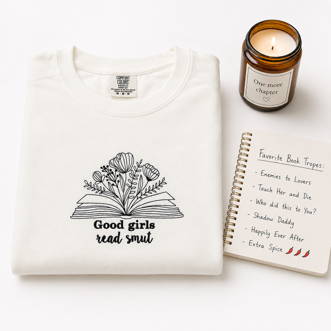 A folded white sweater with a black embroidered graphic of an open book made of flowers and the text 'Good girls read smut' is styled on a white background alongside a brown candle jar and a spiral notebook.