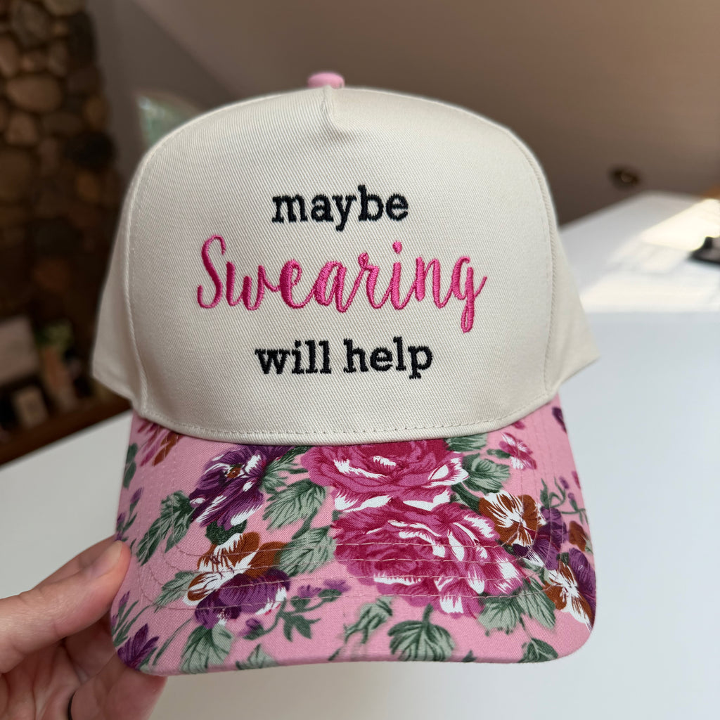 A hand holds a baseball cap with an off-white crown and a pink floral brim. The front of the cap has embroidered text reading 'maybe Swearing will help' in black and pink.