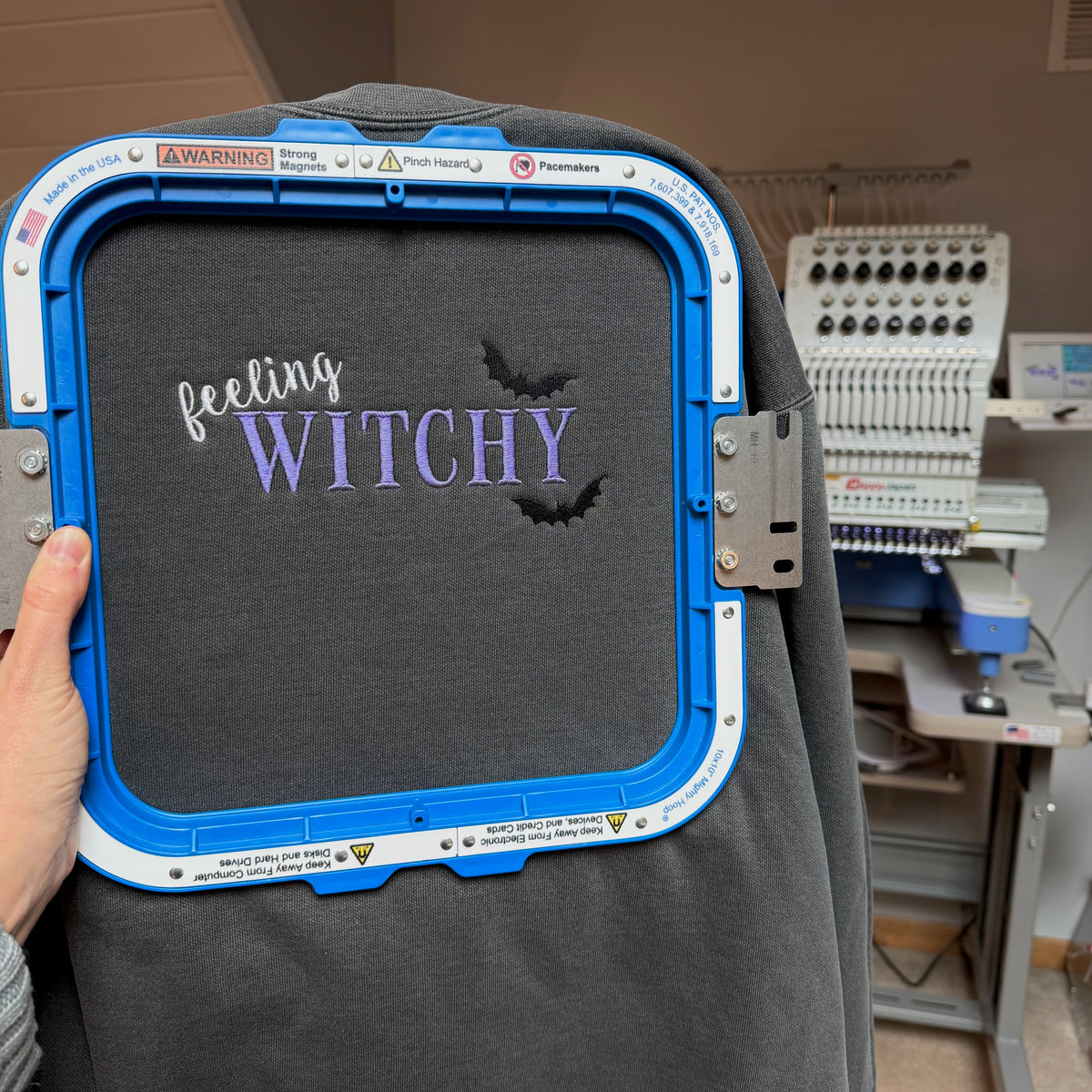 A dark gray sweater inside a blue and white embroidery machine frame, featuring the text 'feeling WITCHY' in white and lavender-purple with two black bats. A hand is holding the frame on the left, and an embroidery machine is visible in the background.