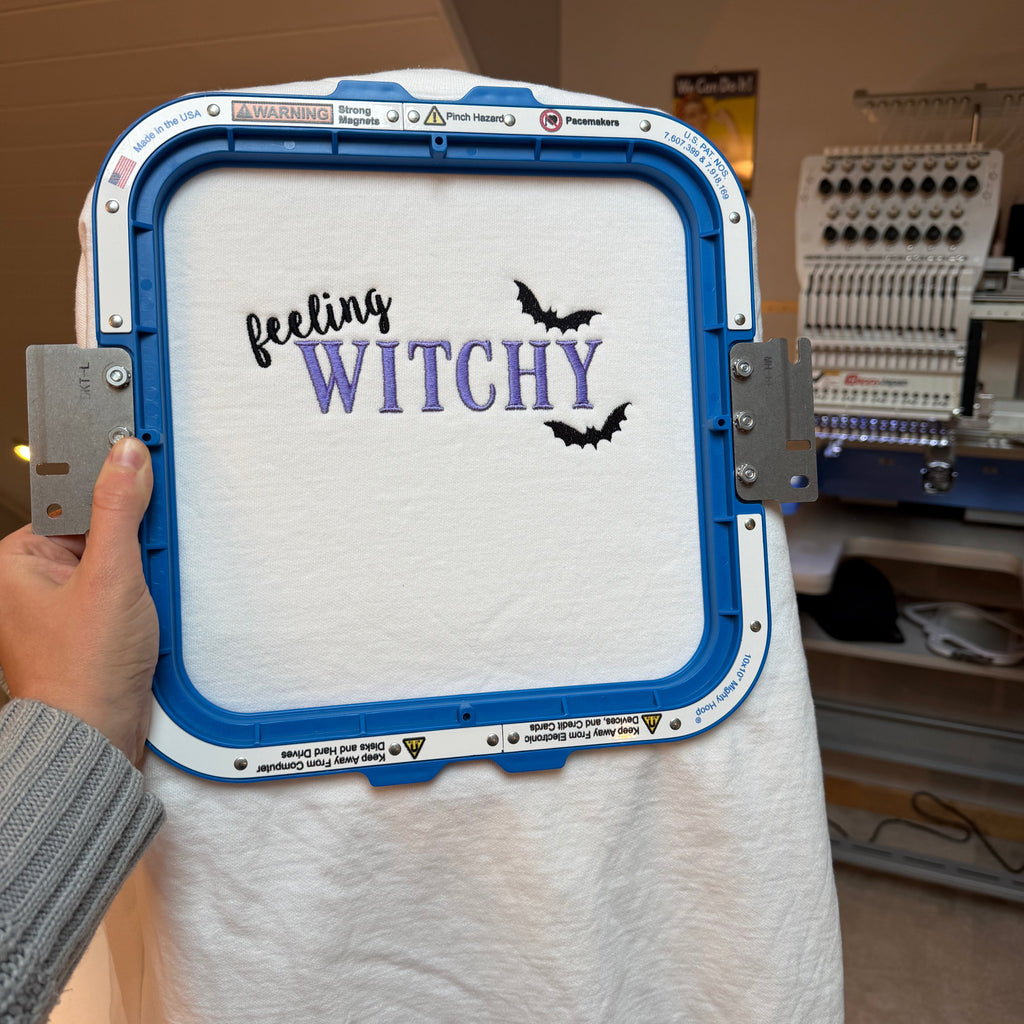 A white sweater held in a blue embroidery hoop by a hand, featuring the text 'feeling WITCHY' in black and purple script with two black bat silhouettes. The background shows an indoor workshop or studio environment with an embroidery machine.