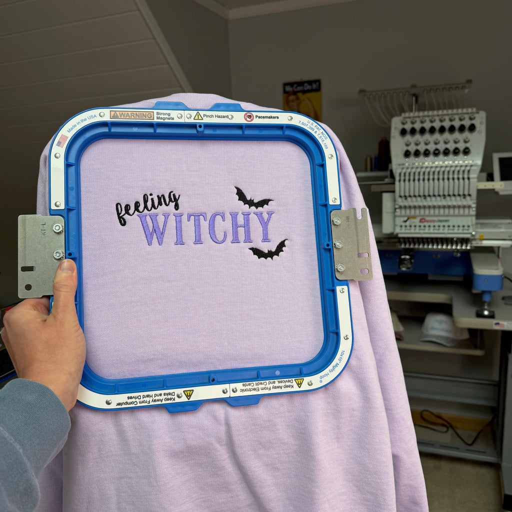 A hand holds an embroidery hoop with a light purple sweater featuring the text 'feeling WITCHY' in black and purple script with two black bat silhouettes. The background shows an embroidery machine in a workshop setting.