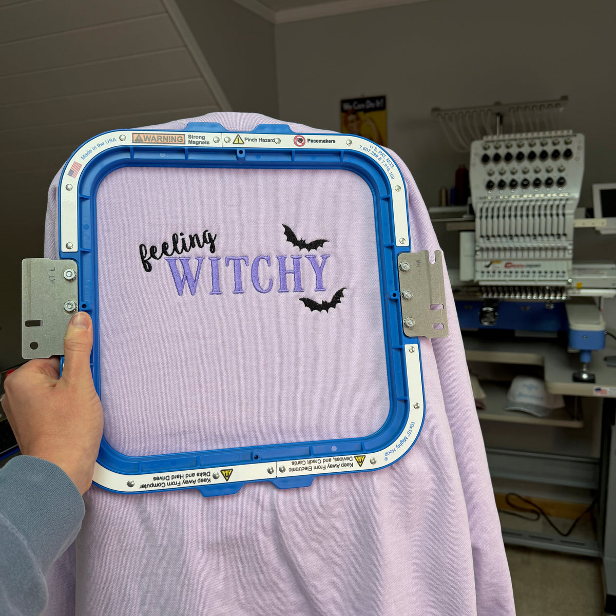 A hand holds an embroidery hoop with a light purple sweater featuring the text 'feeling WITCHY' in black and purple script with two black bat silhouettes. The background shows an embroidery machine in a workshop setting.