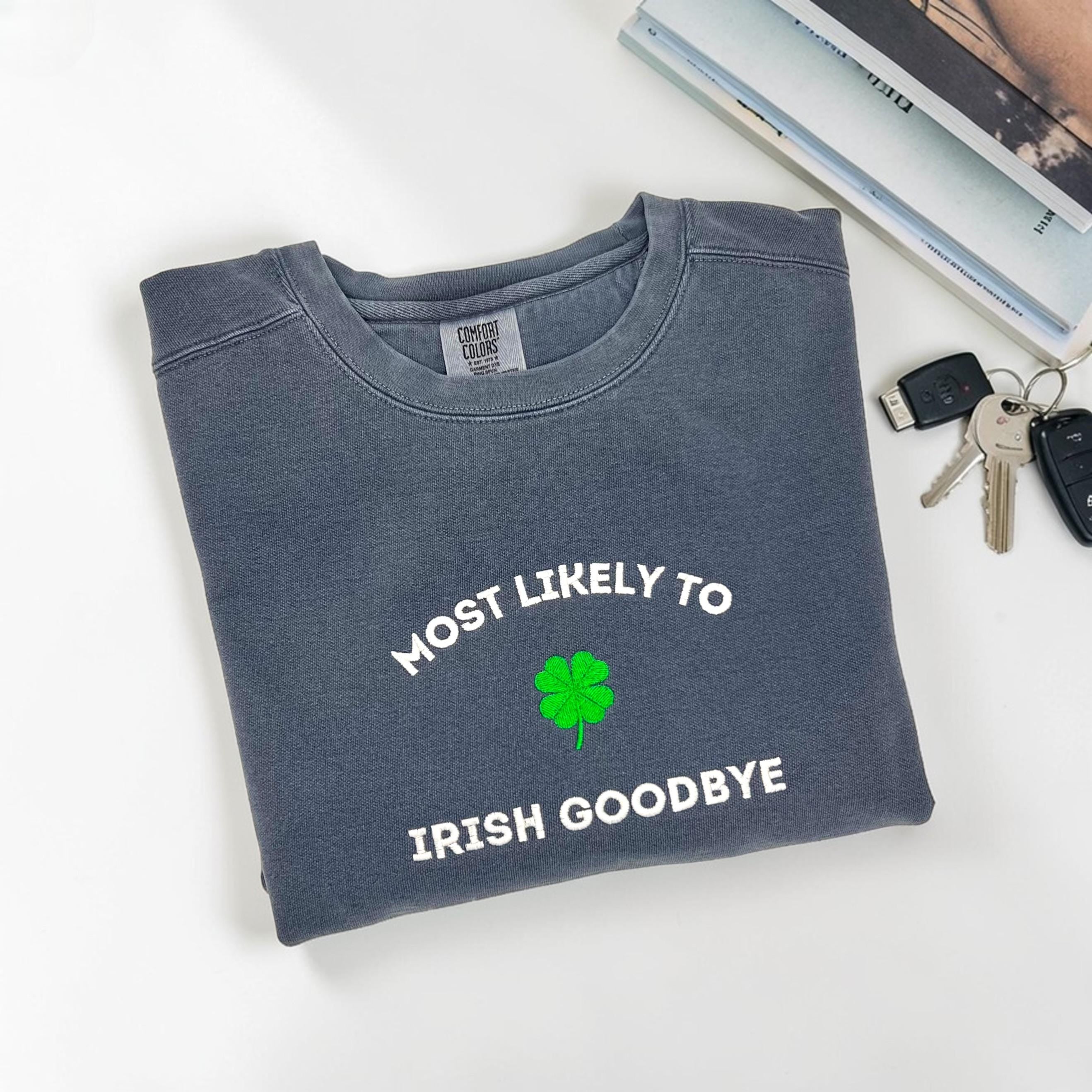 Folded blue sweatshirt with 'MOST LIKELY TO IRISH GOODBYE' and a four leaf clover embroidered on a white surface with keys and books.