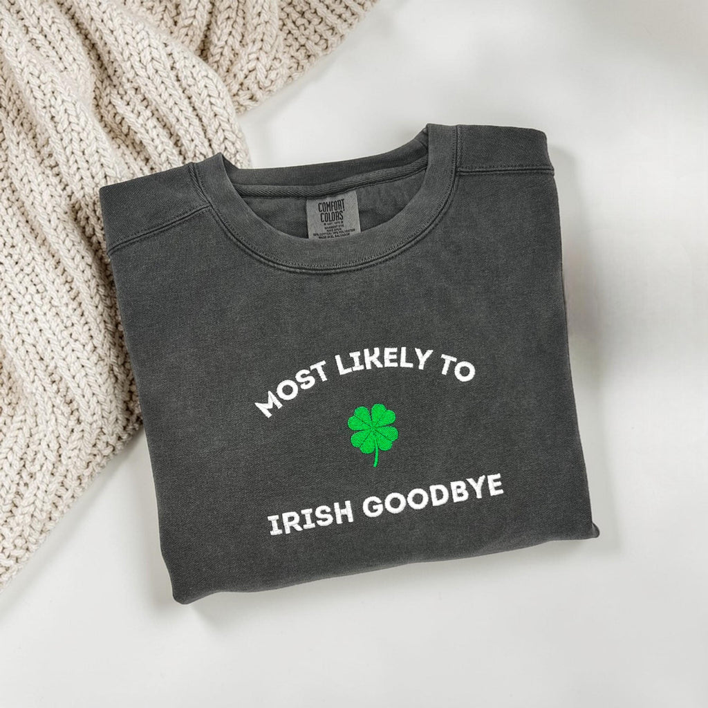 Folded dark grey sweatshirt with 'MOST LIKELY TO IRISH GOODBYE' and a four leaf clover embroidered on a white surface with keys and books.