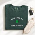A folded dark green crewneck sweatshirt with 'MOST LIKELY TO IRISH GOODBYE' and a green four leaf shamrock graphic embroidered on the front. It is styled in a flat lay with a cream satin sleep mask, a key fob with keys, and soft shadows on a white background.