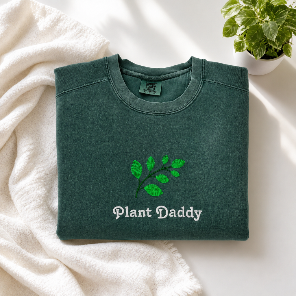 A folded dark green crewneck sweatshirt with an embroidered green leaf graphic and the words 'Plant Daddy' embroidered on the front, styled on a white blanket with a small potted plant in the corner.