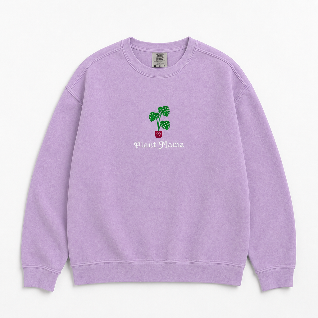 A lavender crewneck sweatshirt featuring an embroidered monstera plant in a pink pot and the text 'Plant Mama' on the chest, displayed against a clean off-white background.