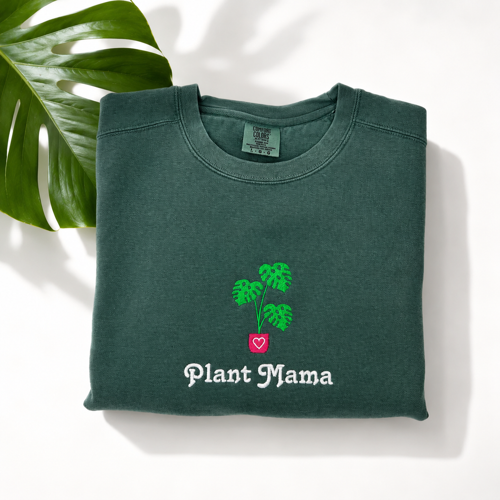 A folded dark green crewneck sweatshirt with an embroidered monstera and the words 'Plant Mama' in white, styled on a white background with a green monstera leaf in the corner.
