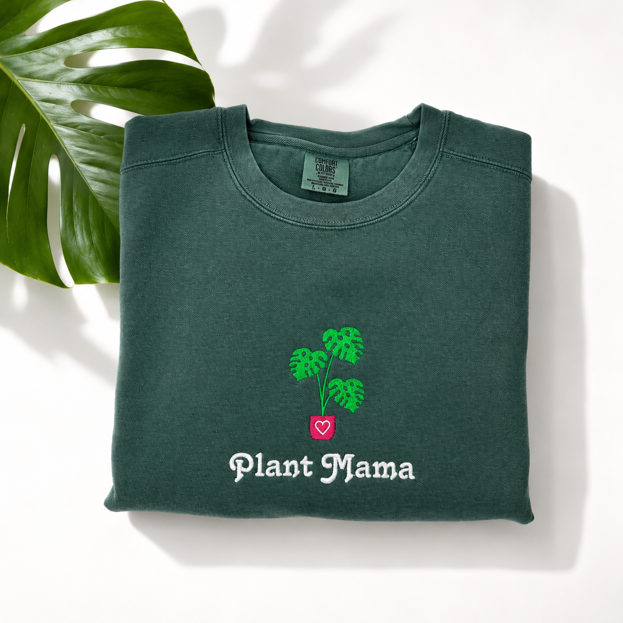 A folded dark green crewneck sweatshirt with an embroidered monstera and the words 'Plant Mama' in white, styled on a white background with a green monstera leaf in the corner.