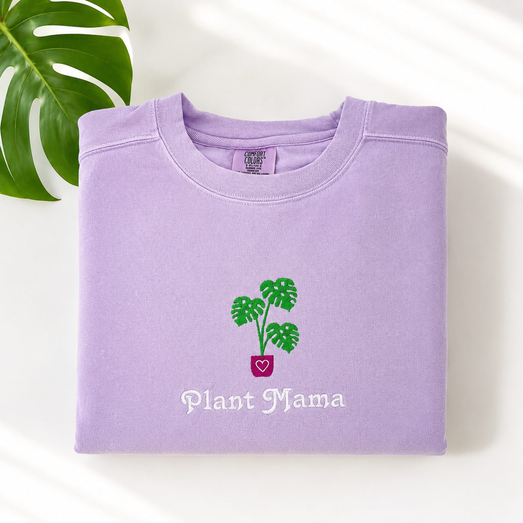 A folded lavender crewneck sweatshirt with an embroidered monstera plant and the words 'Plant Mama' on the front, styled on a white background with a green monstera leaf in the corner.