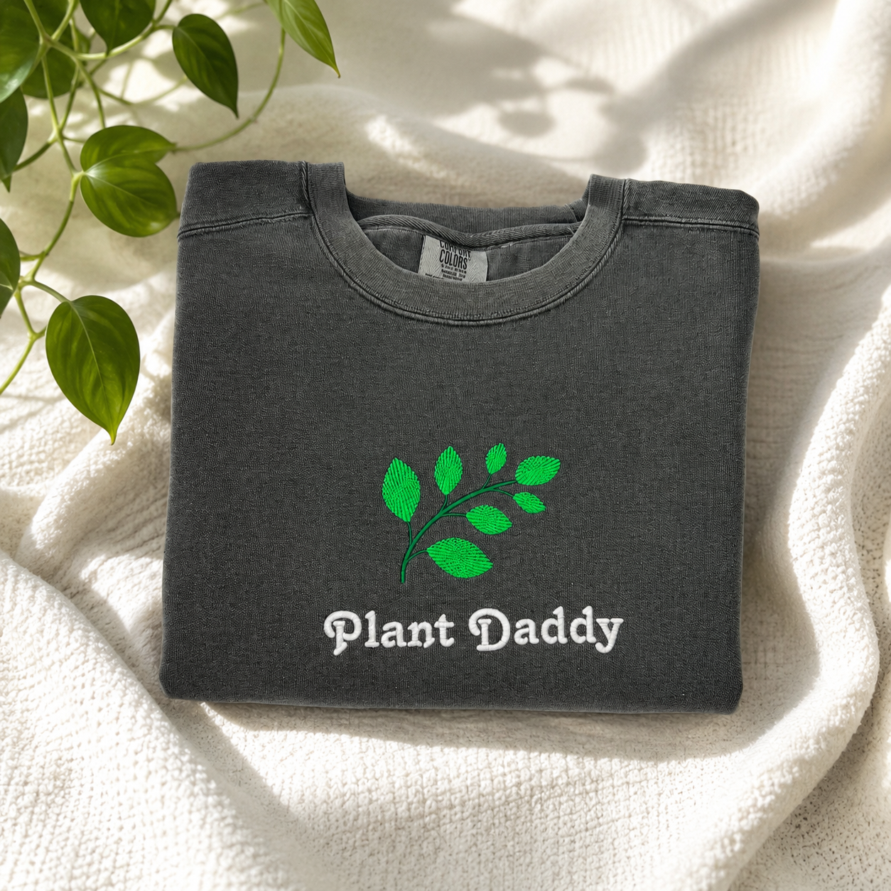 A folded dark gray crewneck sweatshirt with an embroidered green leaf graphic and the words 'Plant Daddy' printed on the front, styled on a textured cream blanket with green plant leaves in the corner.