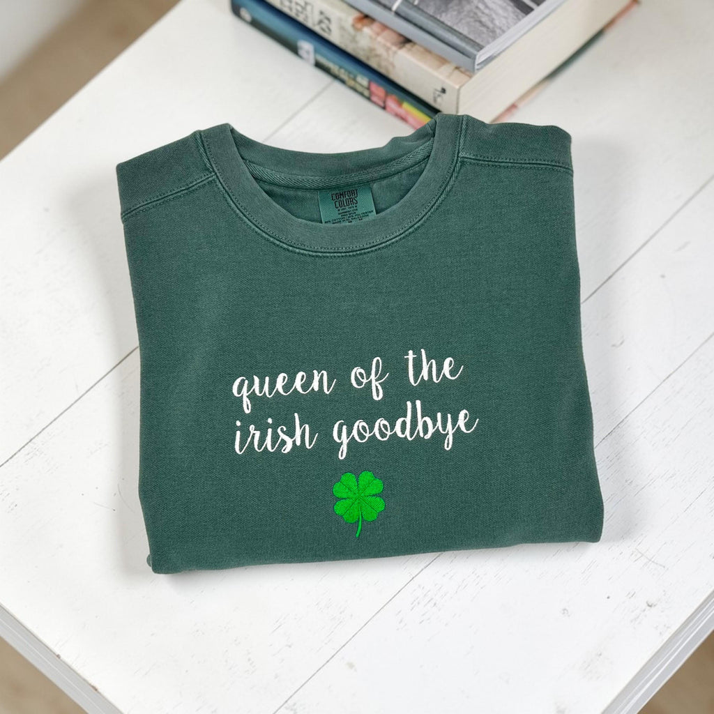 A folded dark green crewneck sweatshirt embroidered with 'queen of the irish goodbye' in white script and a green shamrock, on a white background with a stack of books.