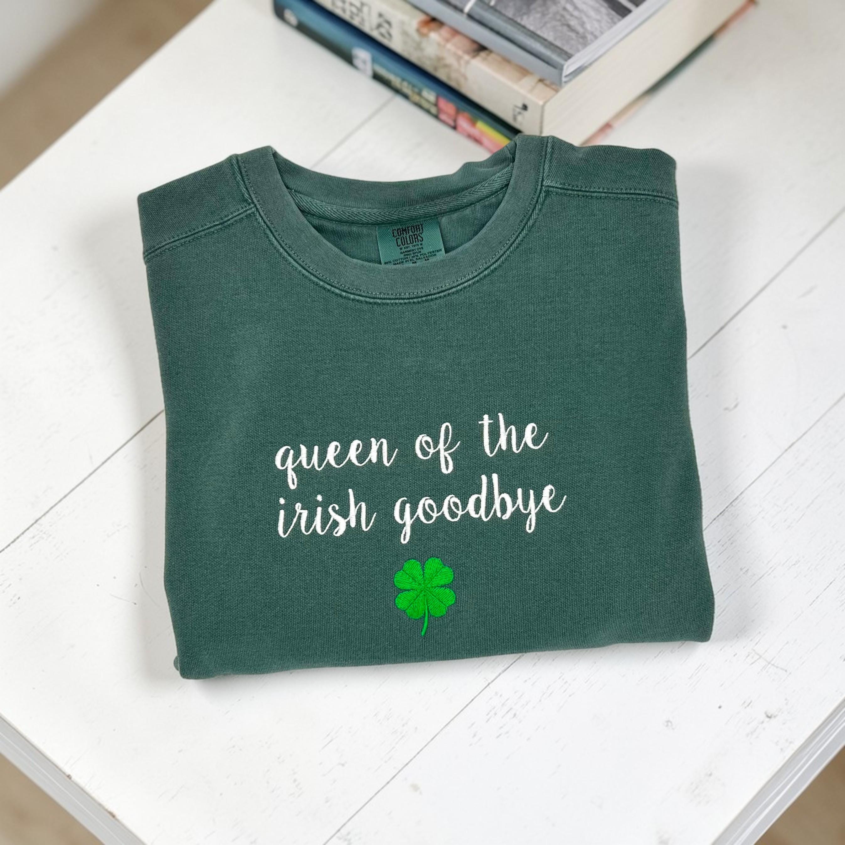 A folded dark green crewneck sweatshirt embroidered with 'queen of the irish goodbye' in white script and a green shamrock, on a white background with a stack of books.
