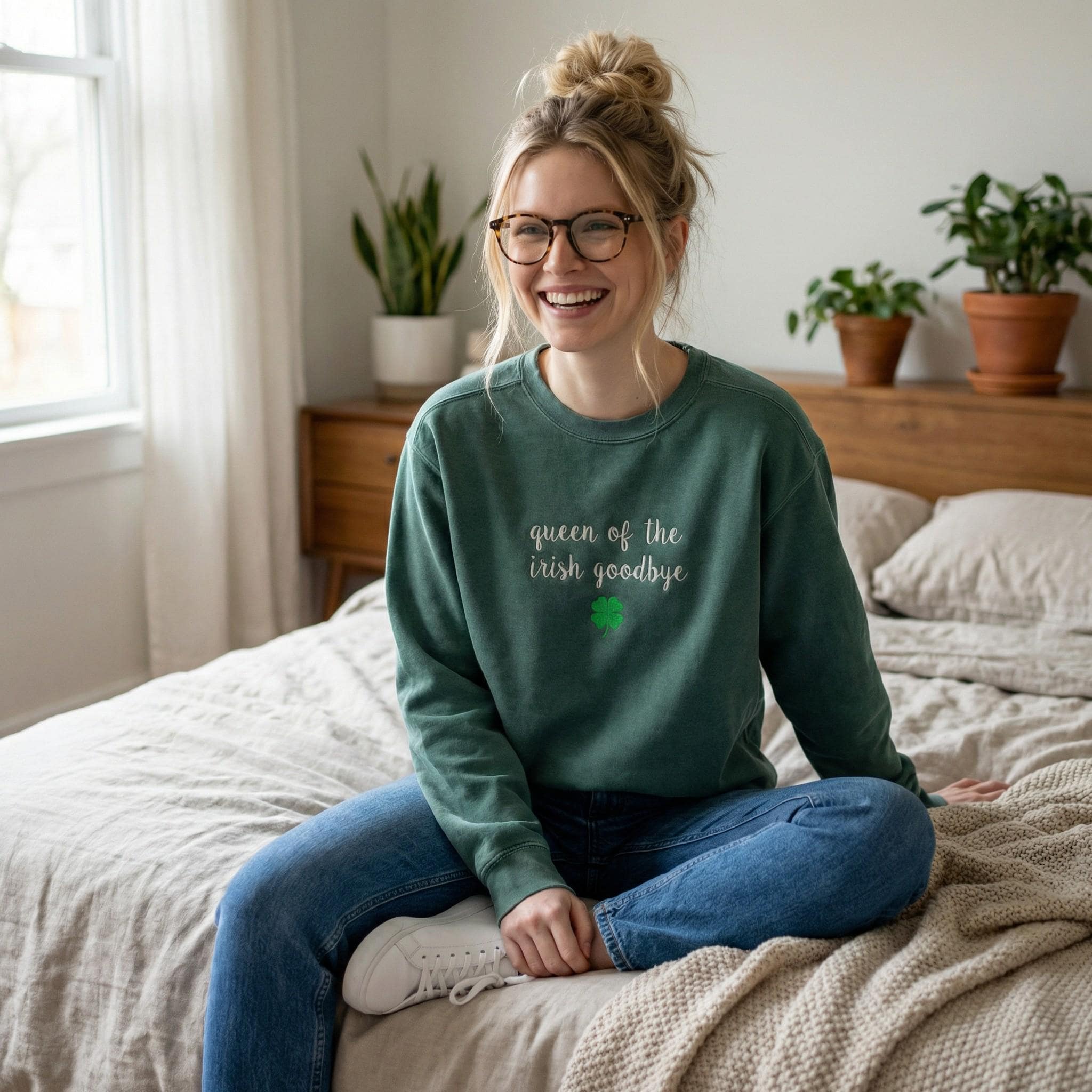 A woman wearing a dark green crewneck sweatshirt embroidered with 'queen of the irish goodbye' in white script and a green shamrock, in a cozy bedroom with plants and books.