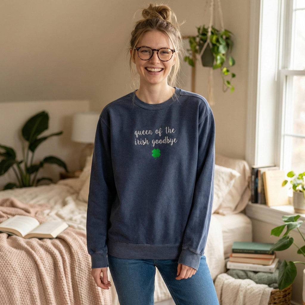 A woman wearing slate blue crewneck sweatshirt embroidered with 'queen of the irish goodbye' in white script and a green shamrock, in a cozy bedroom with plants and books.