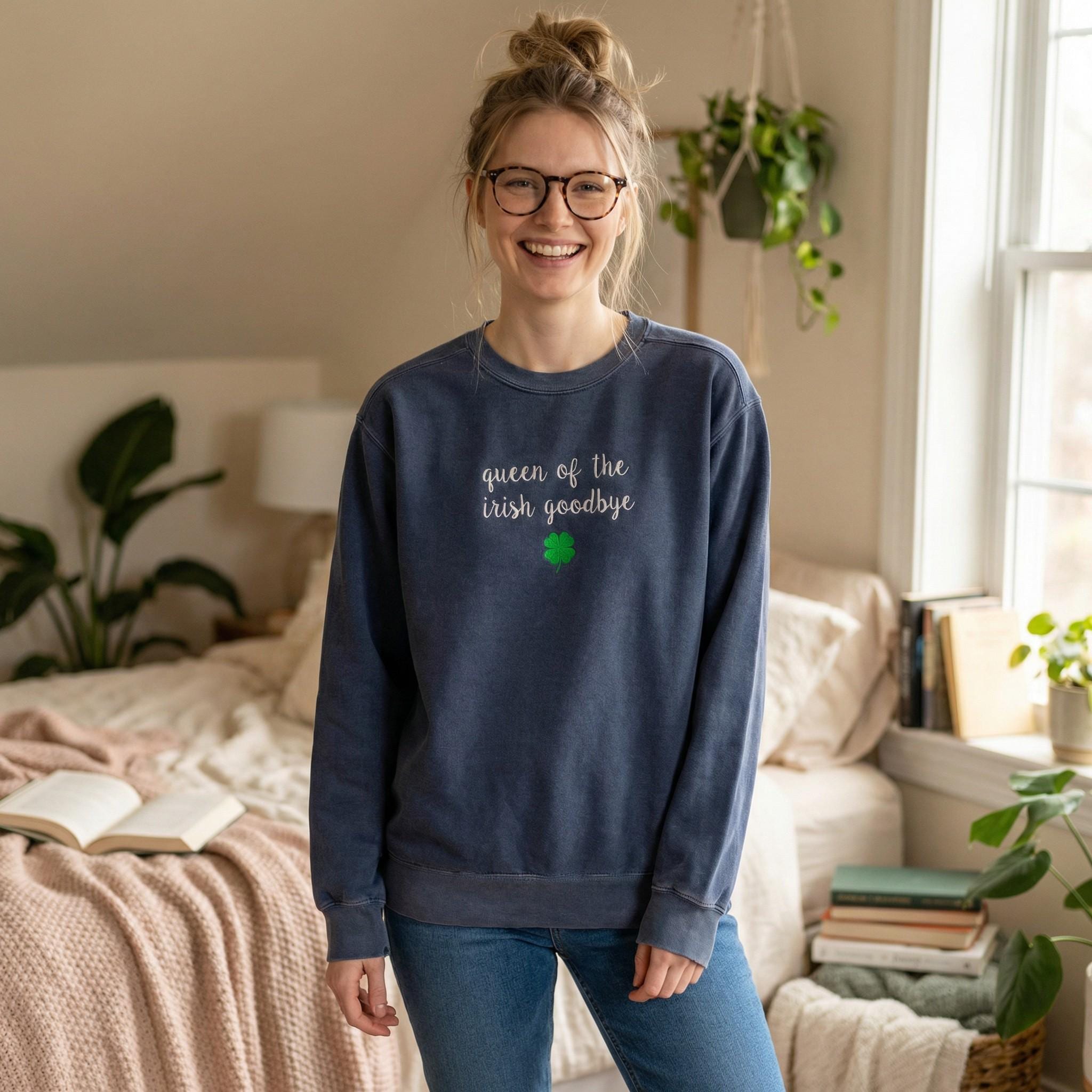 A woman wearing slate blue crewneck sweatshirt embroidered with 'queen of the irish goodbye' in white script and a green shamrock, in a cozy bedroom with plants and books.