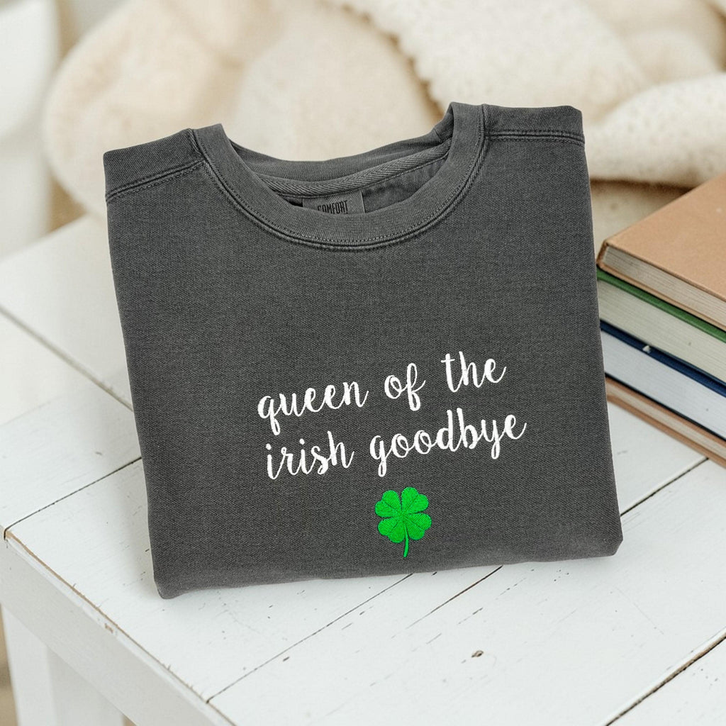 A folded charcoal grey crewneck sweatshirt embroidered with 'queen of the irish goodbye' in white script and a green shamrock, styled in a flat lay with a cream knit blanket, and books on a white table.