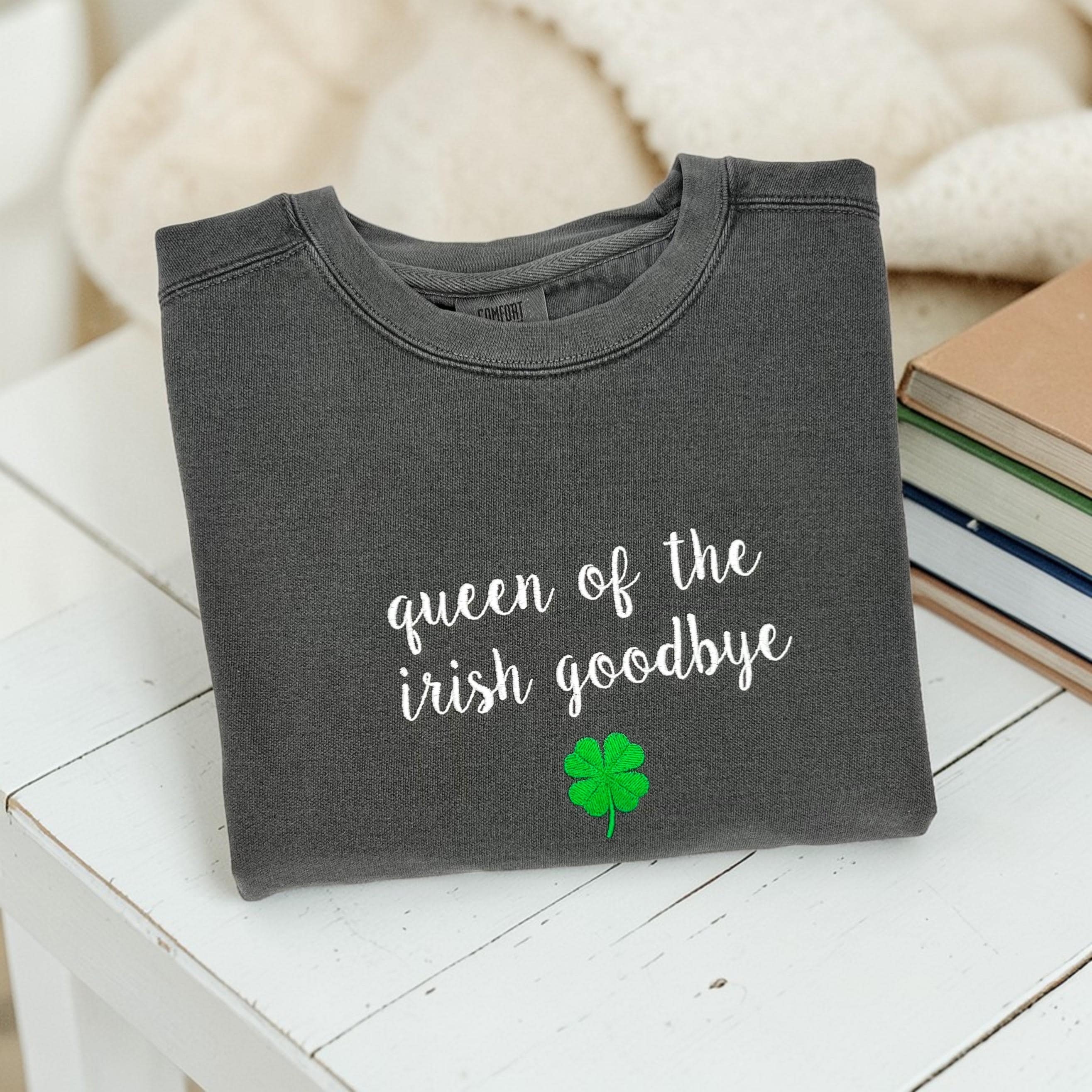 A folded charcoal grey crewneck sweatshirt embroidered with 'queen of the irish goodbye' in white script and a green shamrock, styled in a flat lay with a cream knit blanket, and books on a white table.