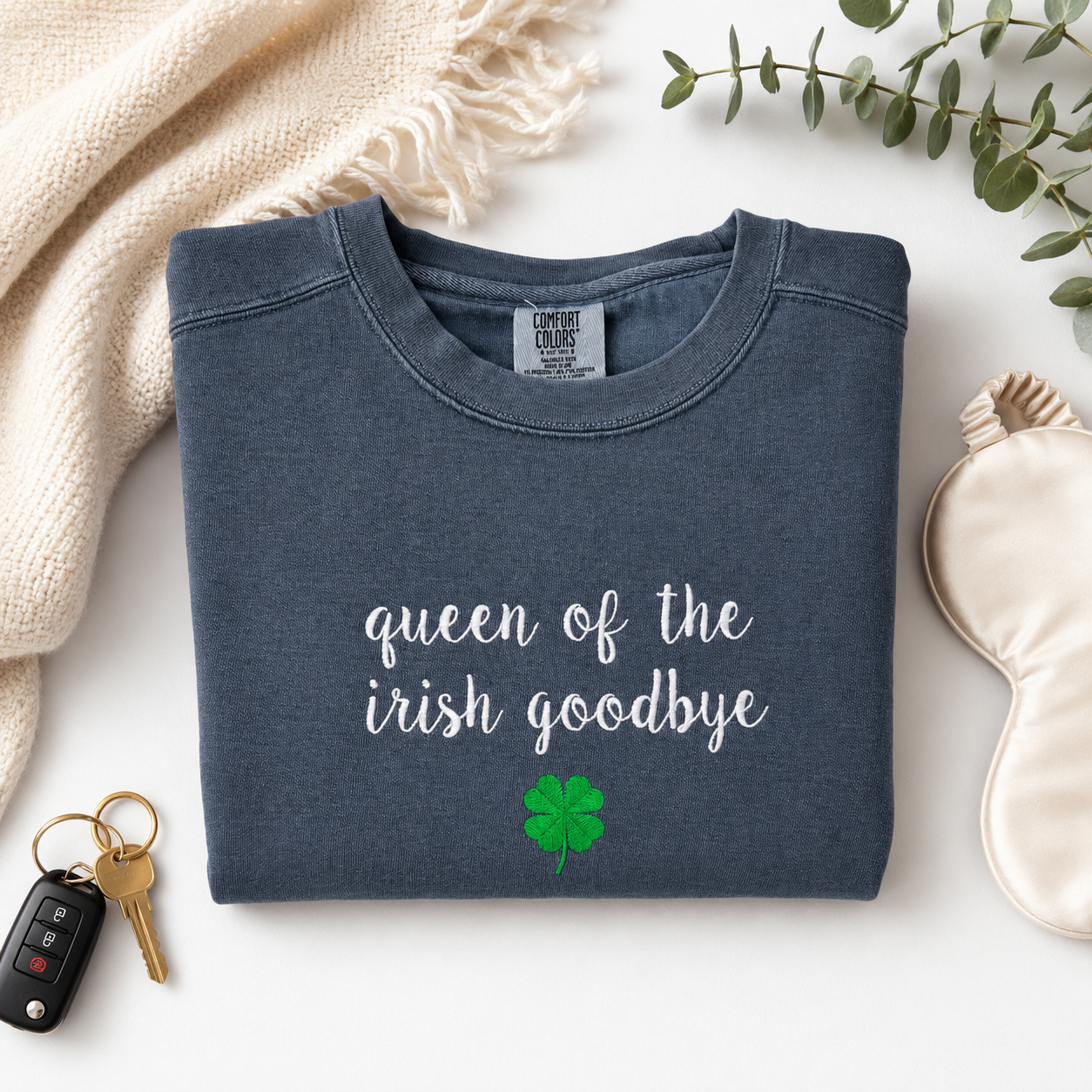 A folded slate blue crewneck sweatshirt embroidered with 'queen of the irish goodbye' in white script and a green shamrock, styled in a flat lay with a cream knit blanket, keys, a sleep mask, and eucalyptus leaves on a white background.