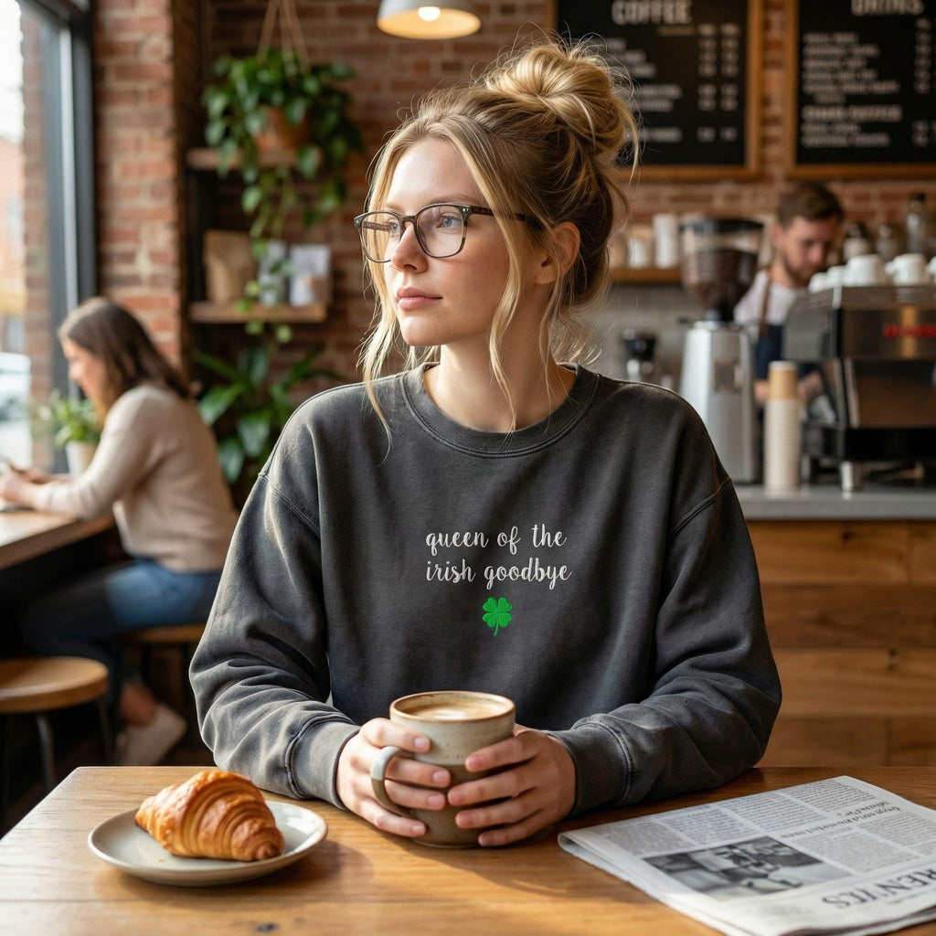 A woman wearing charcoal grey crewneck sweatshirt embroidered with 'queen of the irish goodbye' in white script and a green shamrock, in a cozy cafe with a latte and croissant. 