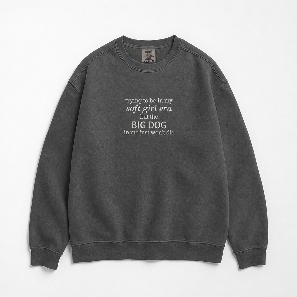 A dark gray crewneck sweatshirt with 'trying to be in my soft girl era but the BIG DOG in me just won't die', displayed flat against a light white background.