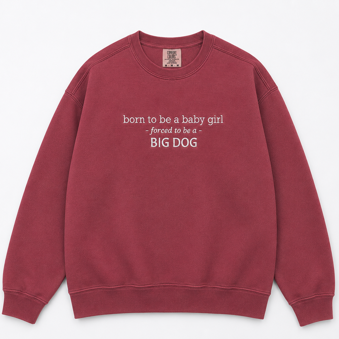 A crewneck sweatshirt in a washed, muted burgundy red, featuring the text 'born to be a baby girl - forced to be a BIG DOG' printed in white on the chest. The sweatshirt is laid flat against a clean, light off-white background.