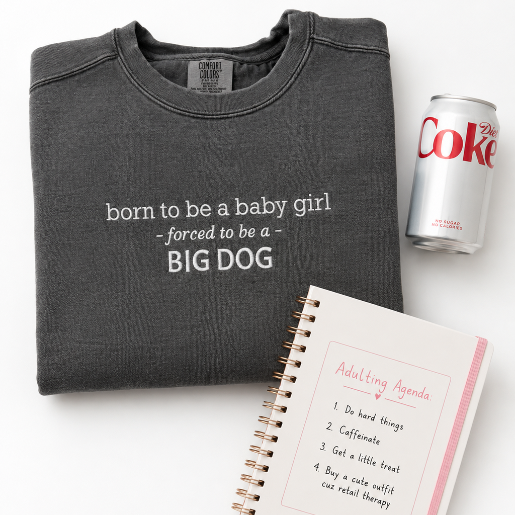A dark charcoal gray crewneck sweatshirt with white text reading 'born to be a baby girl - forced to be a - BIG DOG' is shown in a flat lay with a silver can of Diet Coke and a spiral-bound adulting agenda notebook on a white background.