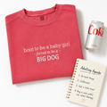 A folded coral pink crewneck sweatshirt with white text embroidered to say 'born to be a baby girl - forced to be a - BIG DOG' is styled in a flat lay with a silver can of Diet Coke and a cream spiral-bound adulting agenda on a white background.