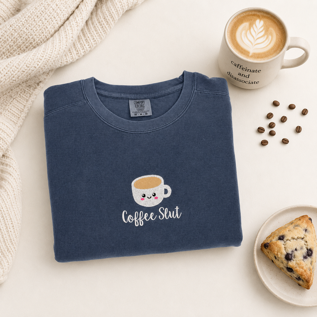 A folded navy blue sweatshirt with a cute coffee cup embroidered and the text 'coffee slut' in white, styled on a light cream background with a coffee mug, coffee beans, a knitted blanket, and a blueberry scone.