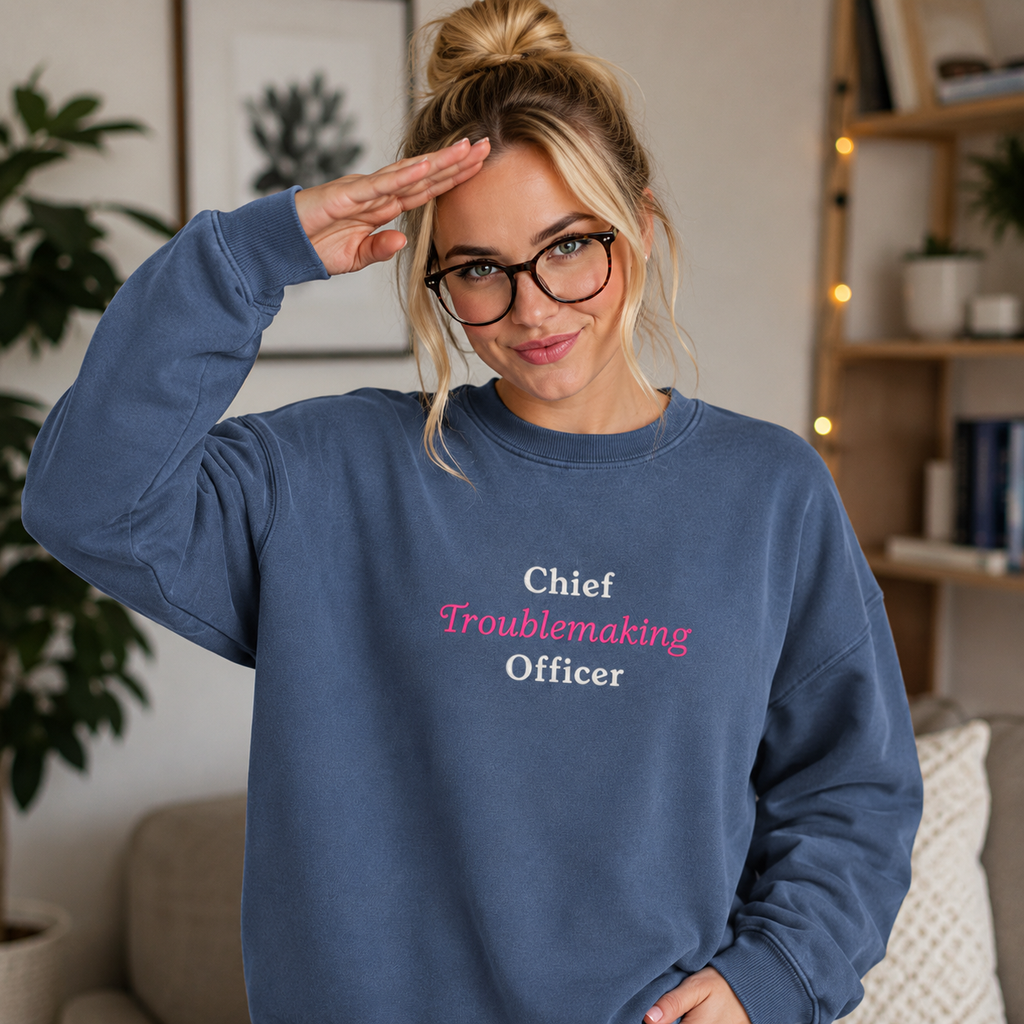 A woman wearing a slate blue crewneck sweatshirt with the text 'Chief Troublemaking Officer' in white and pink, posing with a hand on her head in a cozy indoor setting.