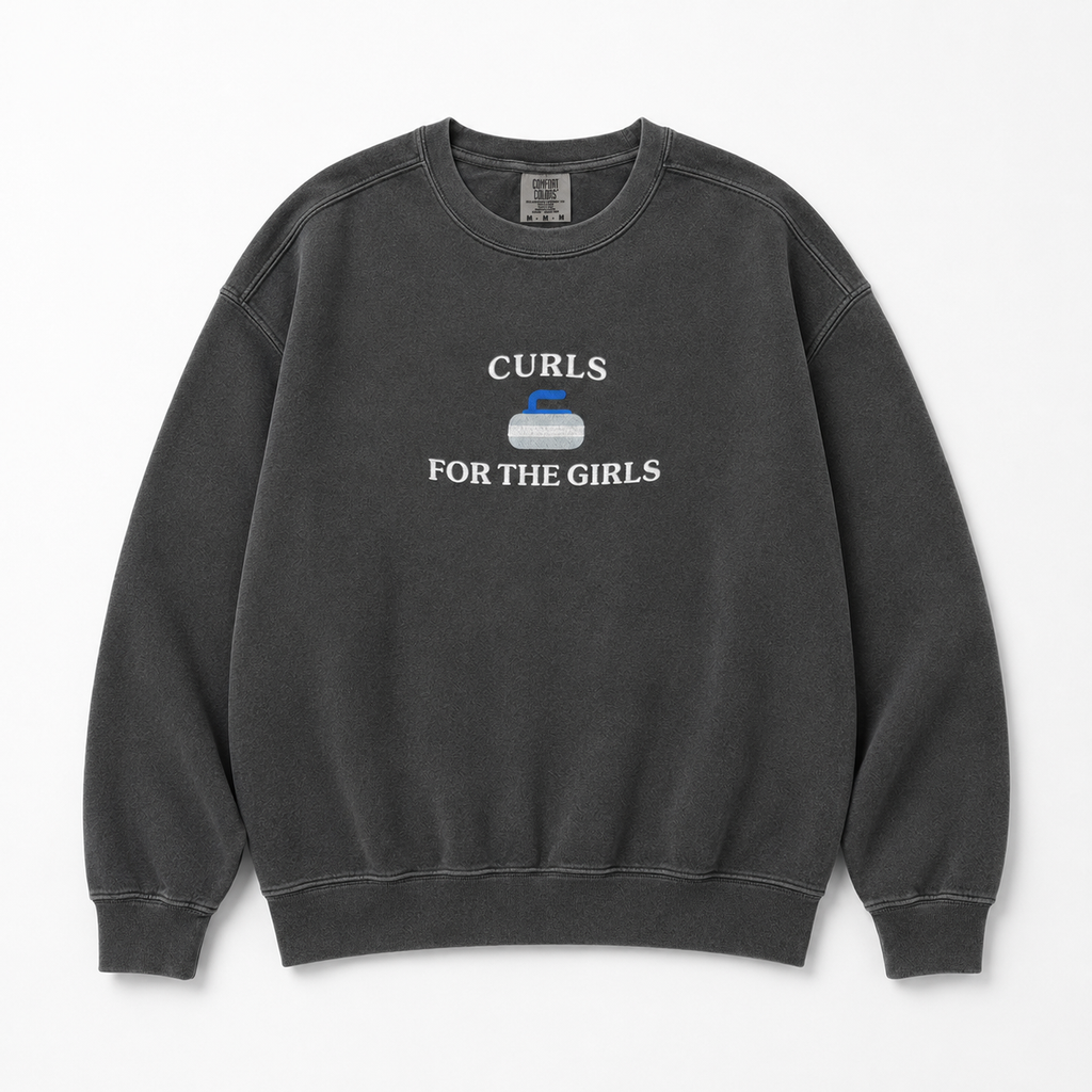 A dark gray washed crewneck sweatshirt featuring a white graphic embroidered that reads 'CURLS FOR THE GIRLS' with a curling stone icon in the center, displayed flat against a light gray background.
