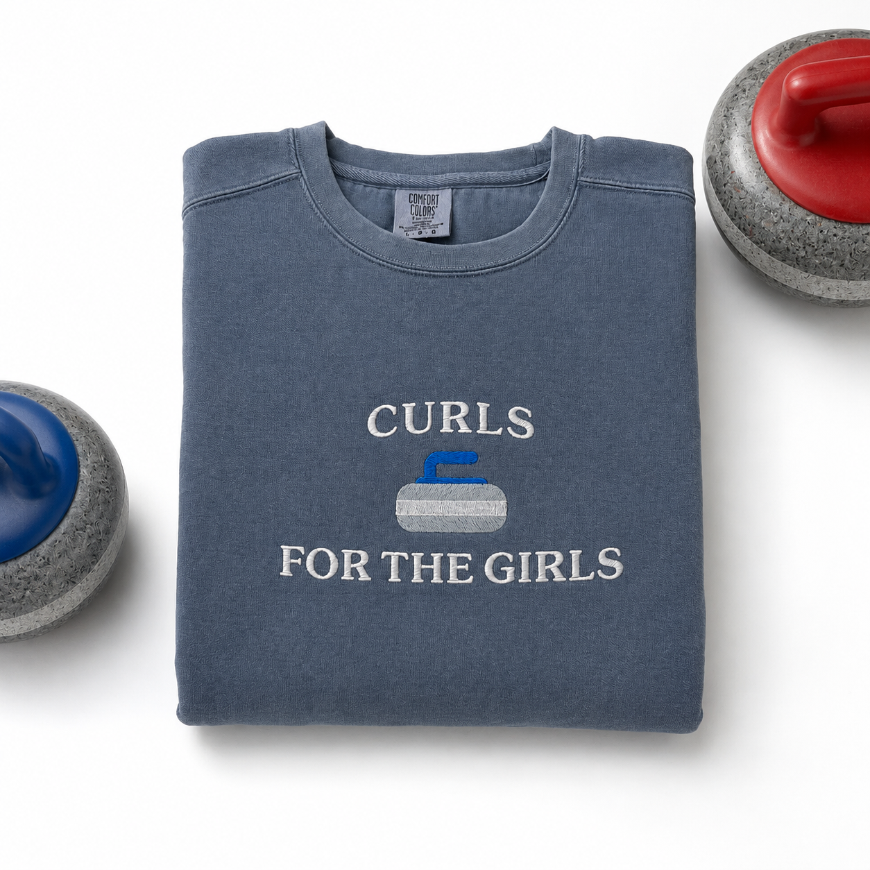 A folded slate blue sweatshirt with the text 'CURLS FOR THE GIRLS' and a curling stone embroidered graphic in white, blue, and gray, displayed on a white background with curling stones partially visible at the corners.