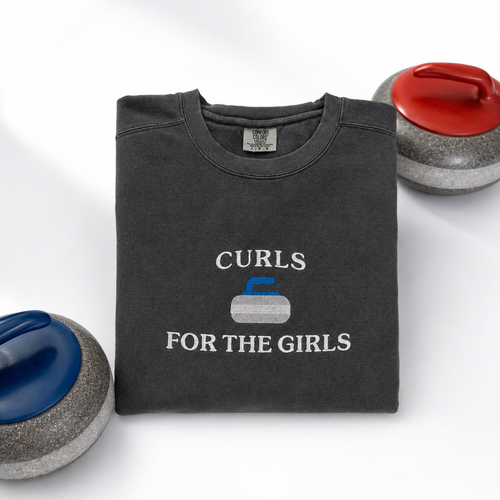 A folded dark charcoal gray sweatshirt with the text 'CURLS FOR THE GIRLS' and a curling stone embroidered graphic, styled on a white background with curling stones partially visible at the corners.
