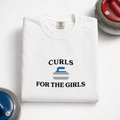 A folded white sweatshirt on a white background with 'CURLS FOR THE GIRLS' text and a curling stone graphic embroidered on the front. Curling stones are partially visible in the corners as styling props.