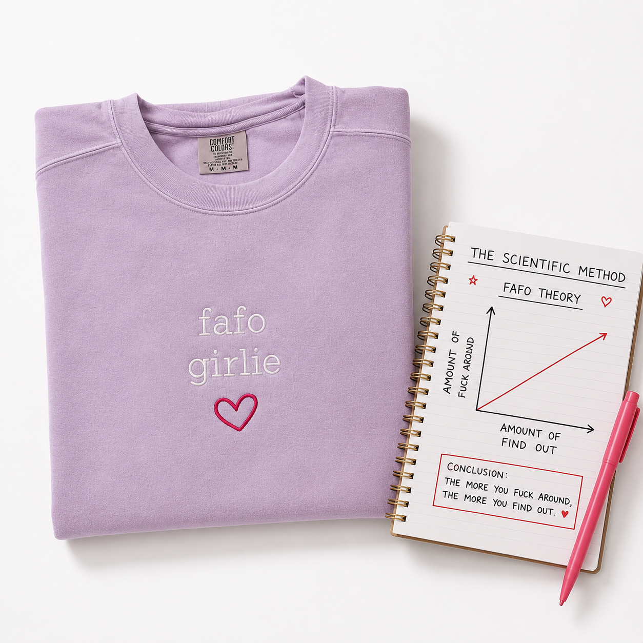 A folded light purple sweatshirt with 'fafo girlie' and a pink heart embroidered on the front, styled on a white background next to a spiral notebook and a pink pen.