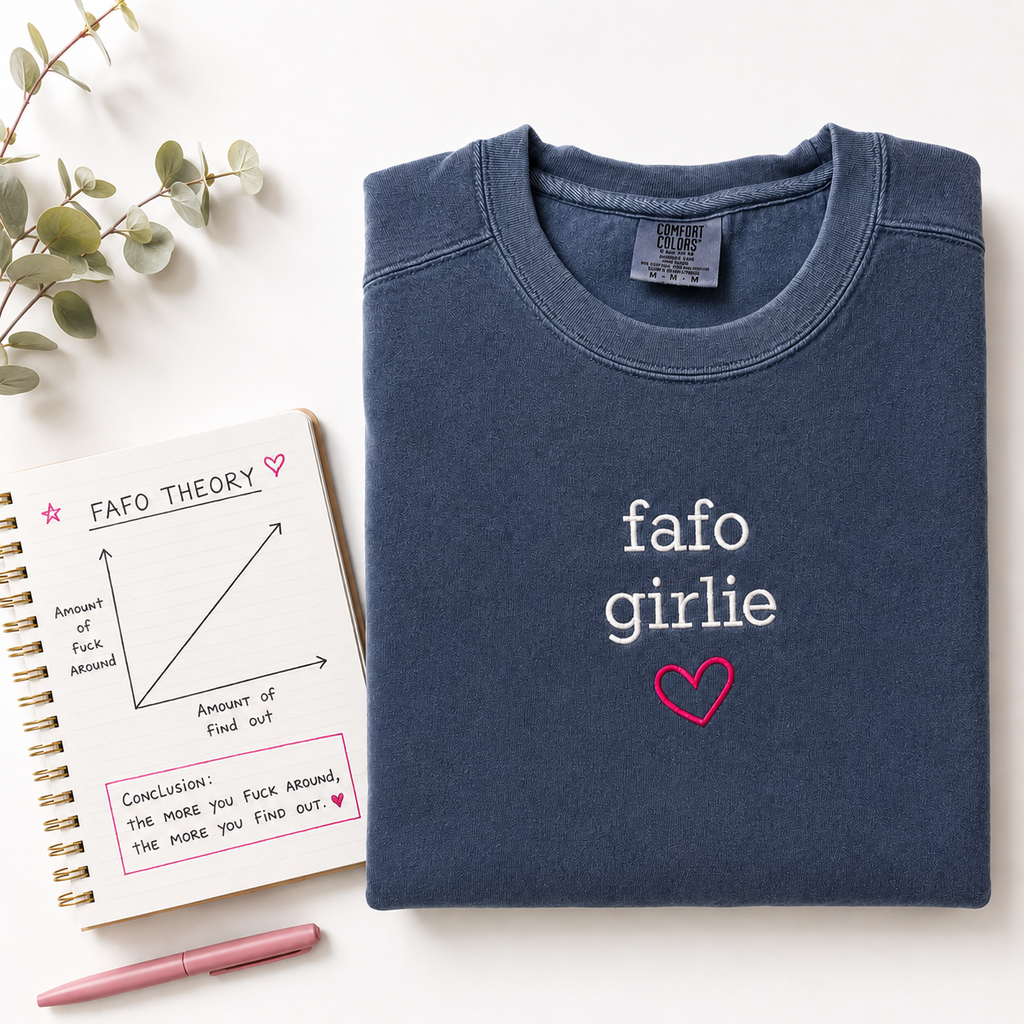 A folded navy blue sweatshirt with 'fafo girlie' in white and a pink heart embroidered on the front, styled on a white background with a spiral notebook, eucalyptus branch, and pink pen.