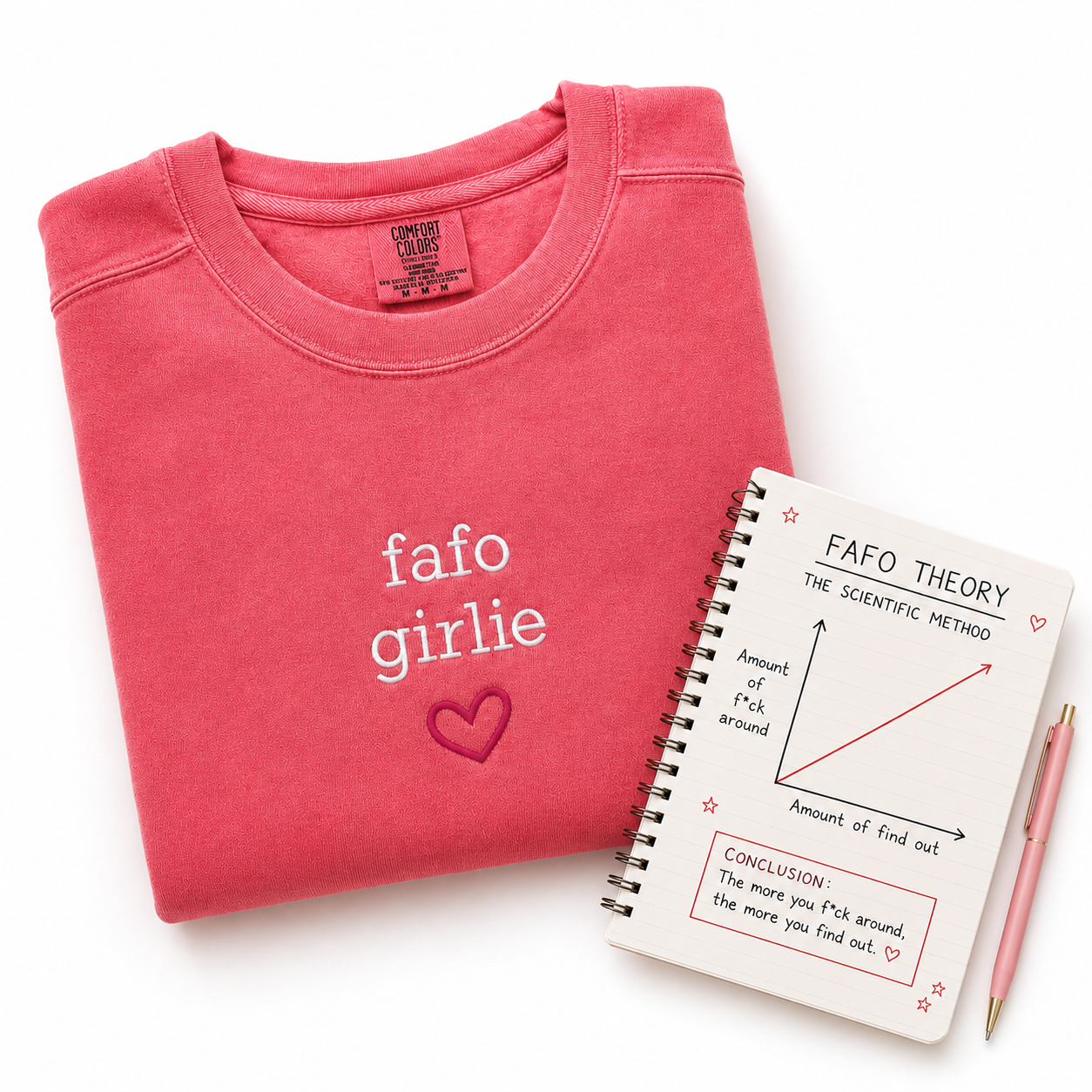 A folded pink sweatshirt with 'fafo girlie' and a heart graphic printed on the front, styled on a white background next to a spiral notebook titled 'FAFO THEORY' and a pink pen.